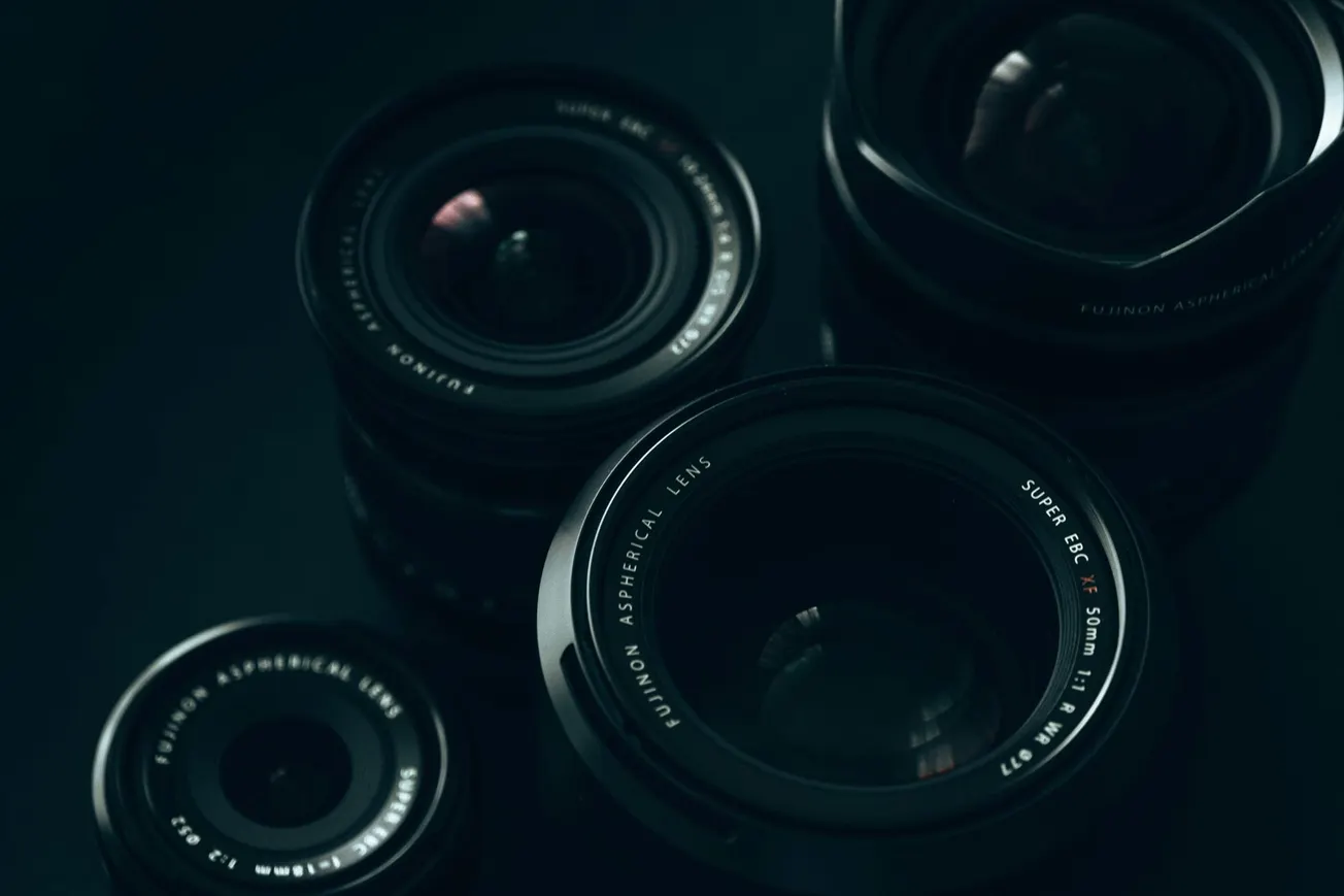 Close-up photo of four camera lenses arranged on a dark surface. The lenses vary in size, displaying brand details.