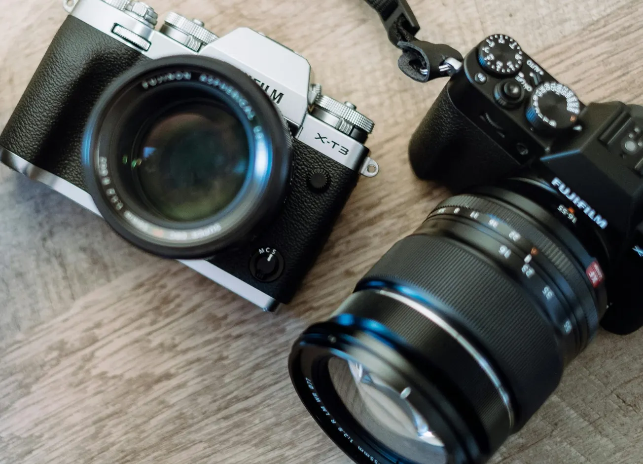 Close-up of two black and silver cameras with large lenses on a wooden surface, angled toward each other.