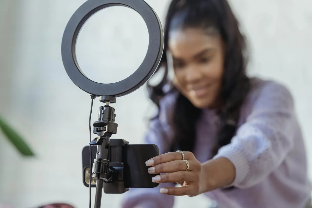A woman in a purple sweater sets up her smartphone on a tripod with a ring light, preparing for a video recording or livestream.