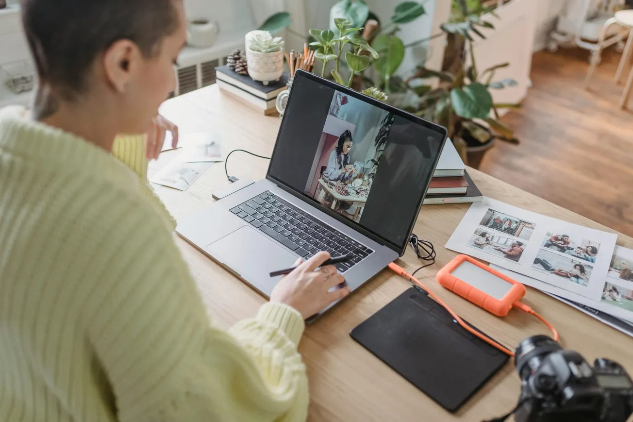 A person in a yellow sweater works at a wooden desk using a laptop, with a photo editing program open. The workspace is decorated with plants.