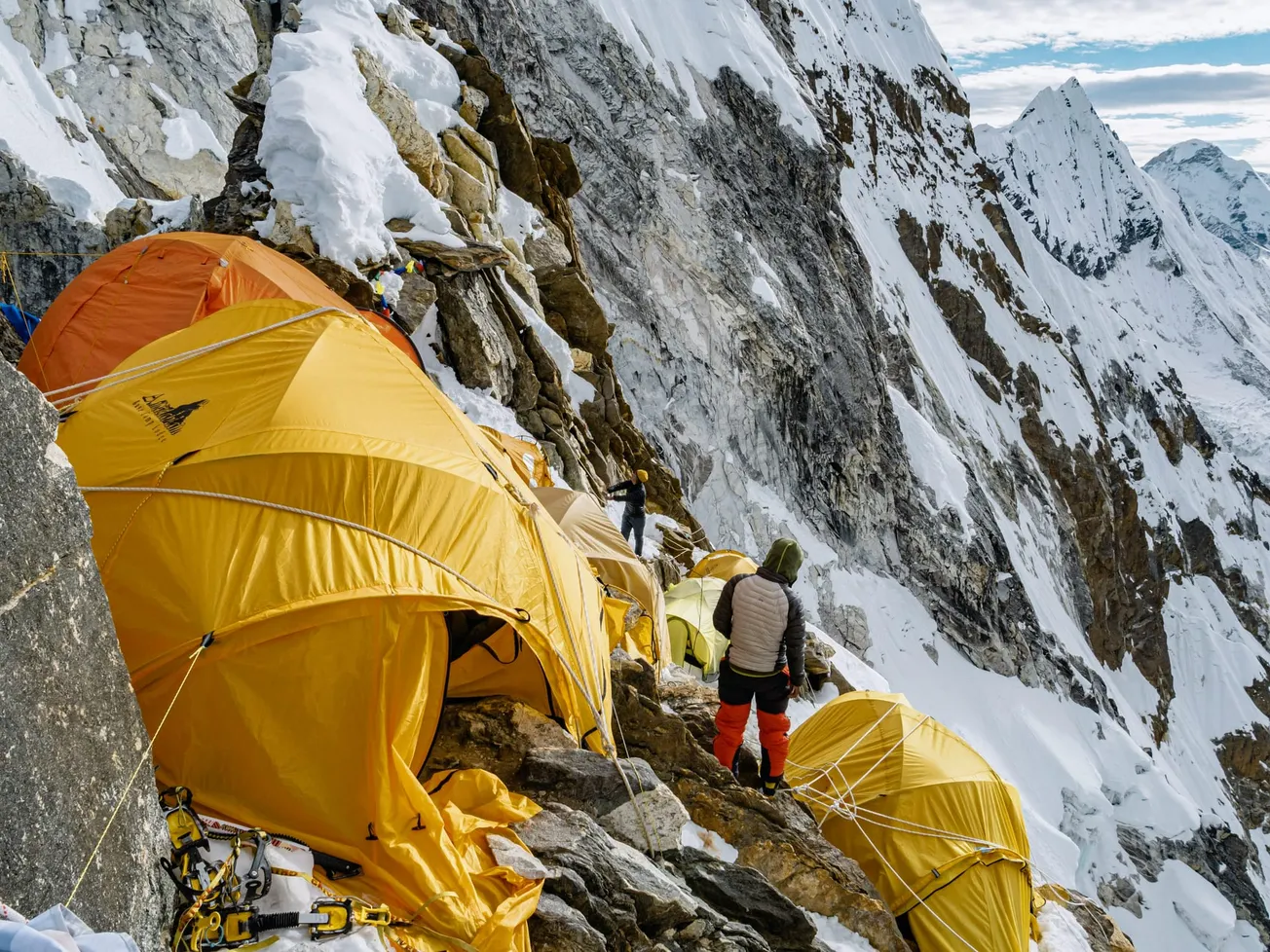 Mountain climbers set up bright yellow tents on a steep, snowy slope. The scene displays harsh, challenging conditions.