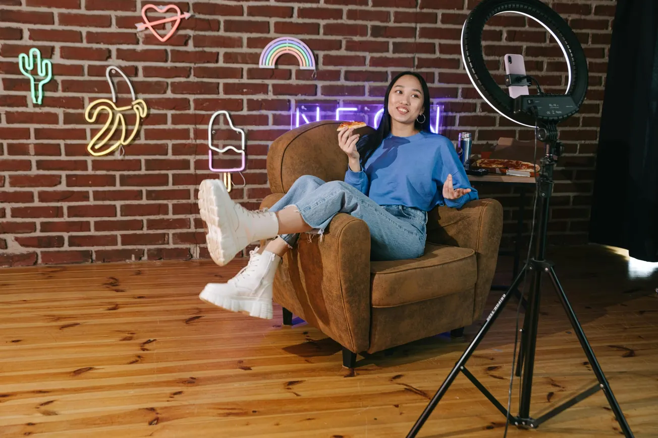 Young woman in a blue sweater sits in a brown chair, holding pizza, in front of a brick wall with neon lights. A ring light and phone are set up.