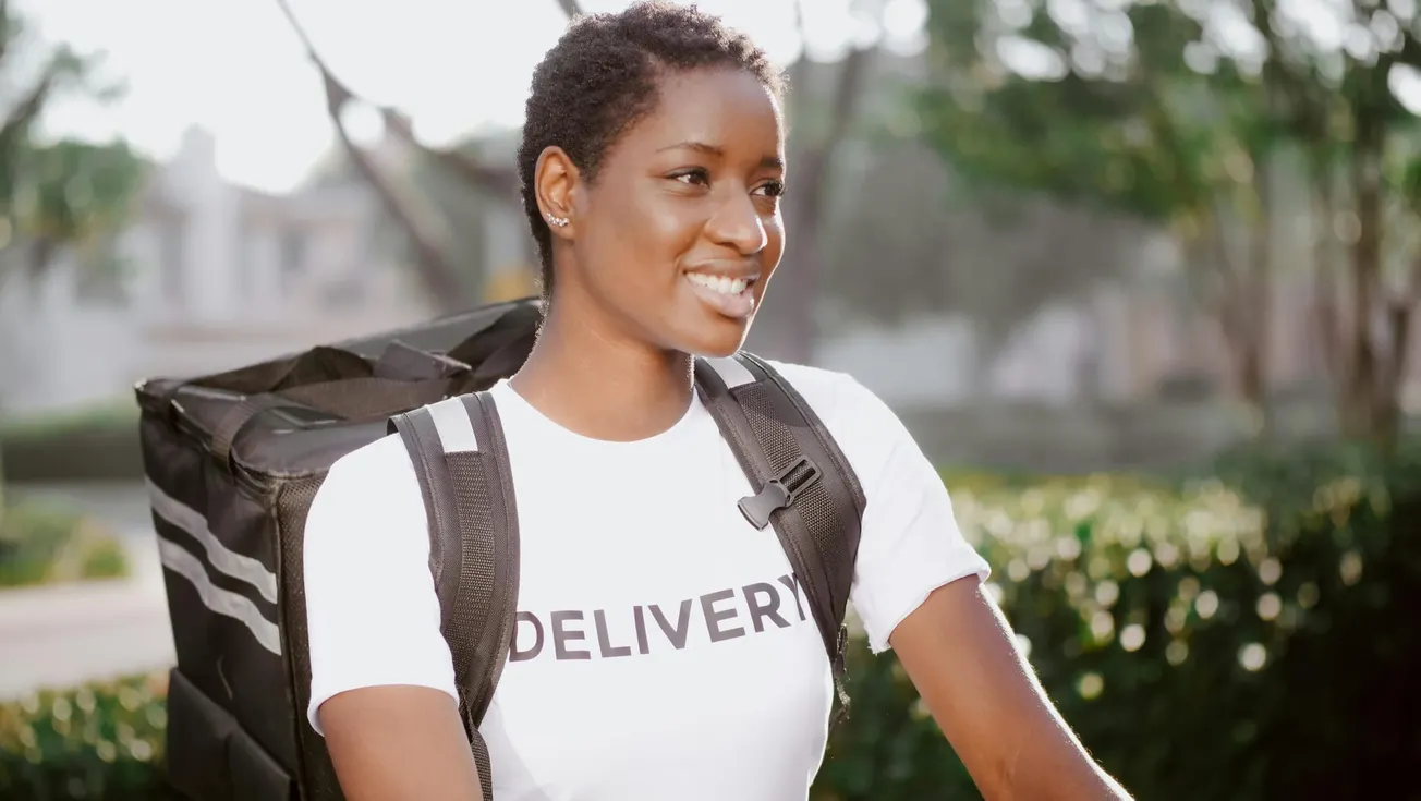 A person wearing a white T-shirt with "DELIVERY" printed on it and a large backpack smiles while outdoors. The background is bright and leafy.