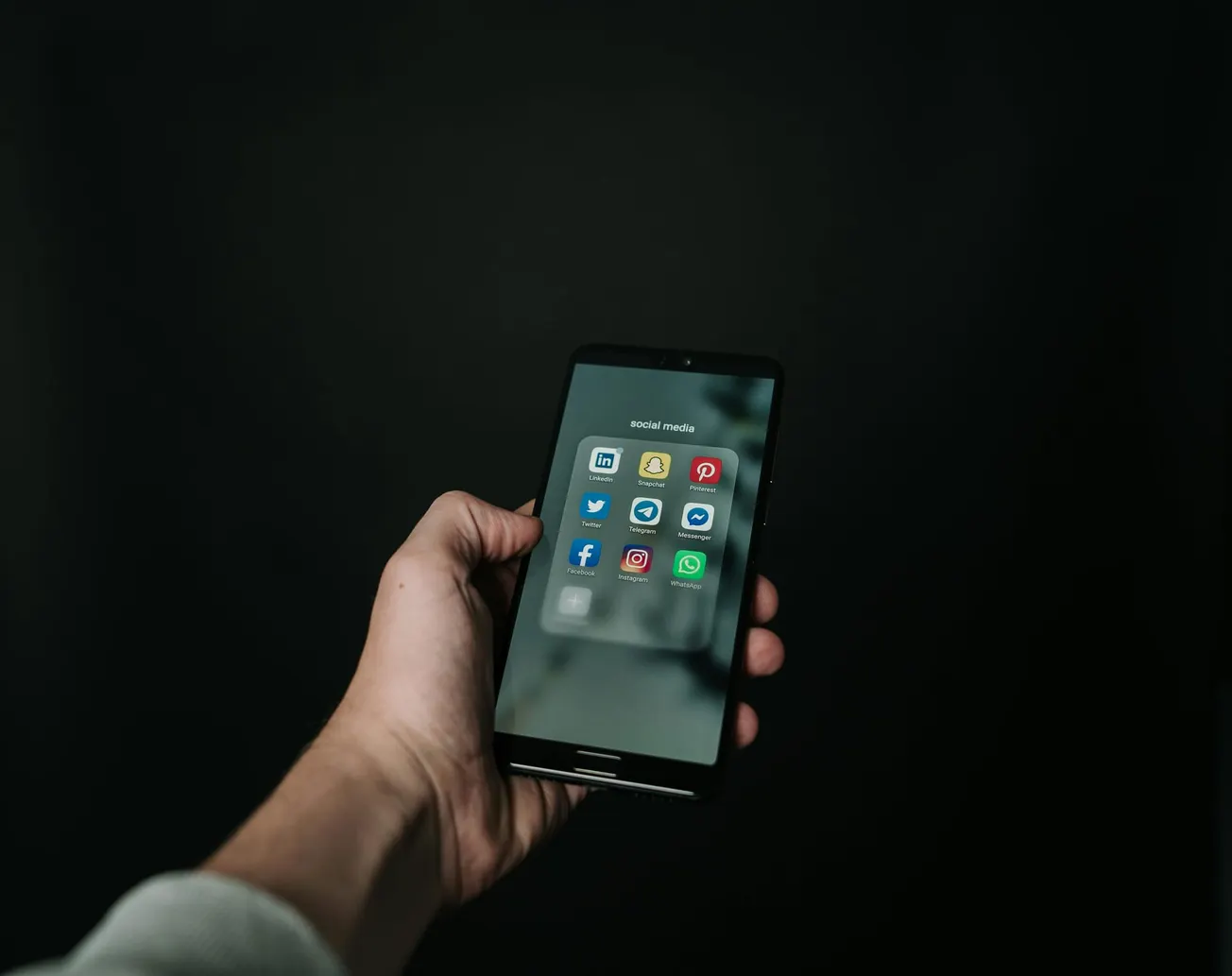 A hand holds a smartphone displaying social media app icons on a dark background.