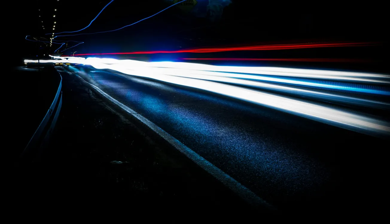 Blurred light trails of white, blue, and red from fast-moving vehicles on a dark road at night.