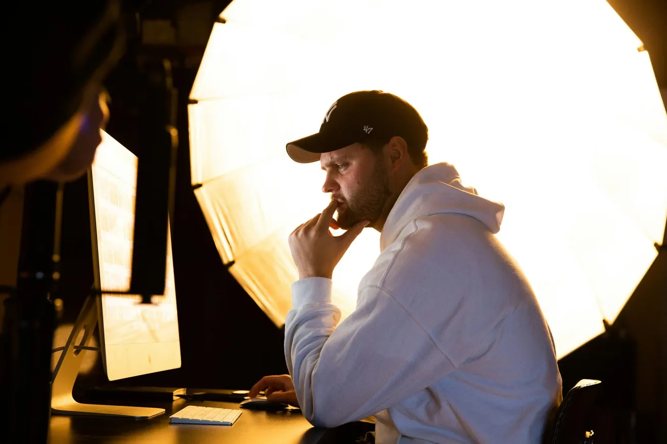 A person in a white hoodie and black cap sits thoughtfully at a computer, illuminated by a large, bright light source.