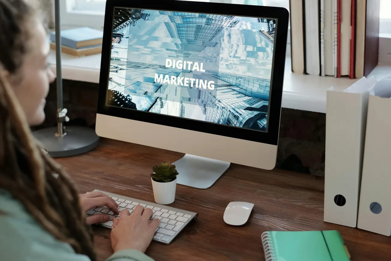 A person at a wooden desk, typing on a computer displaying "Digital Marketing." Nearby are a mouse, potted plant, books, and folders.