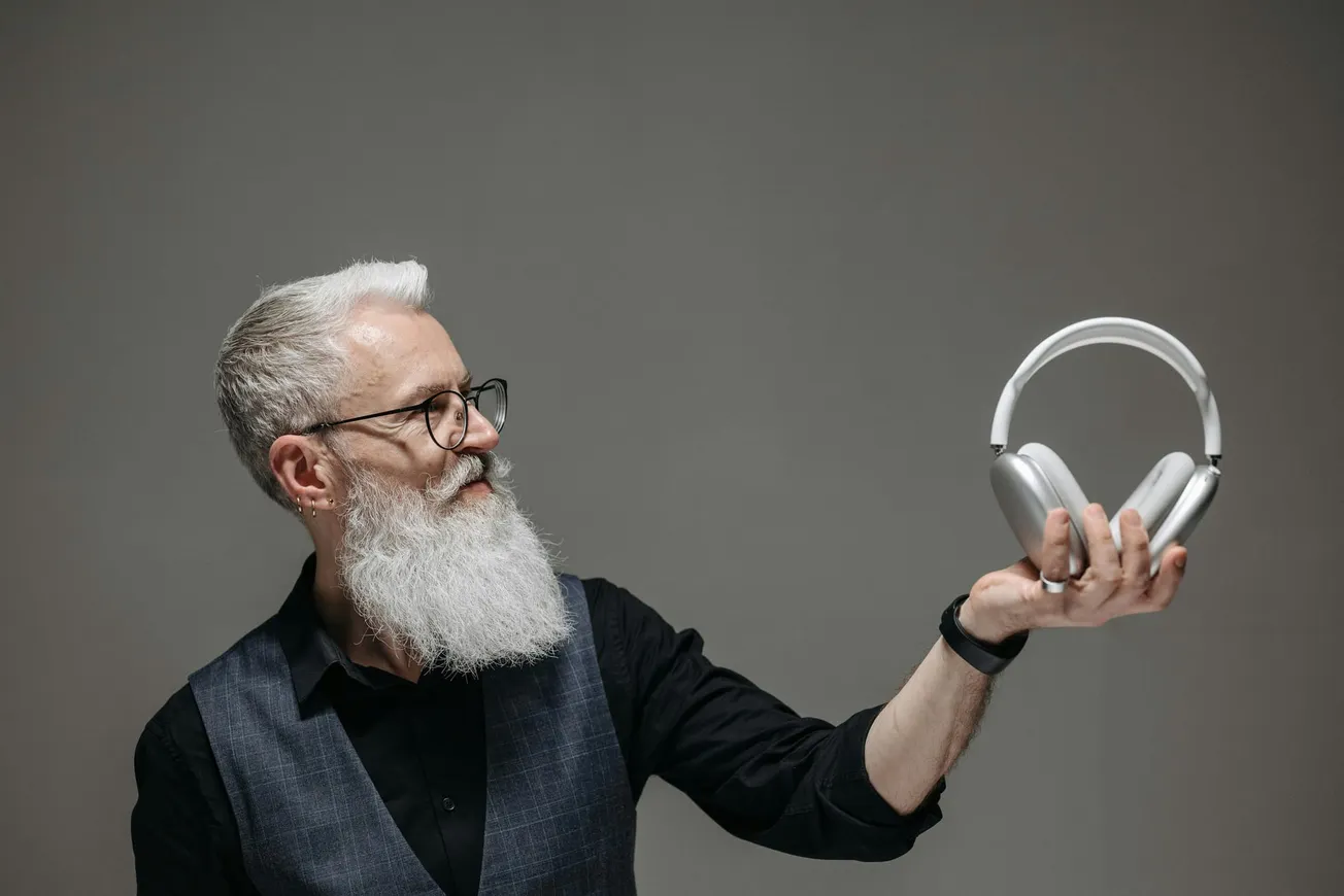 A bearded man with glasses and a vest holds silver headphones, looking at them thoughtfully against a plain gray background.