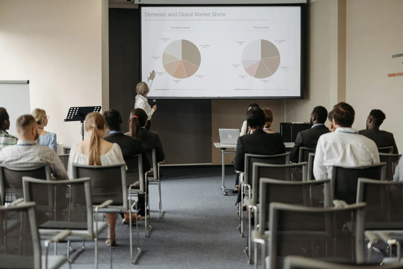 A presenter points to pie charts titled "Domestic and Global Market Share" on a screen in a conference room. Attendees sit attentively.