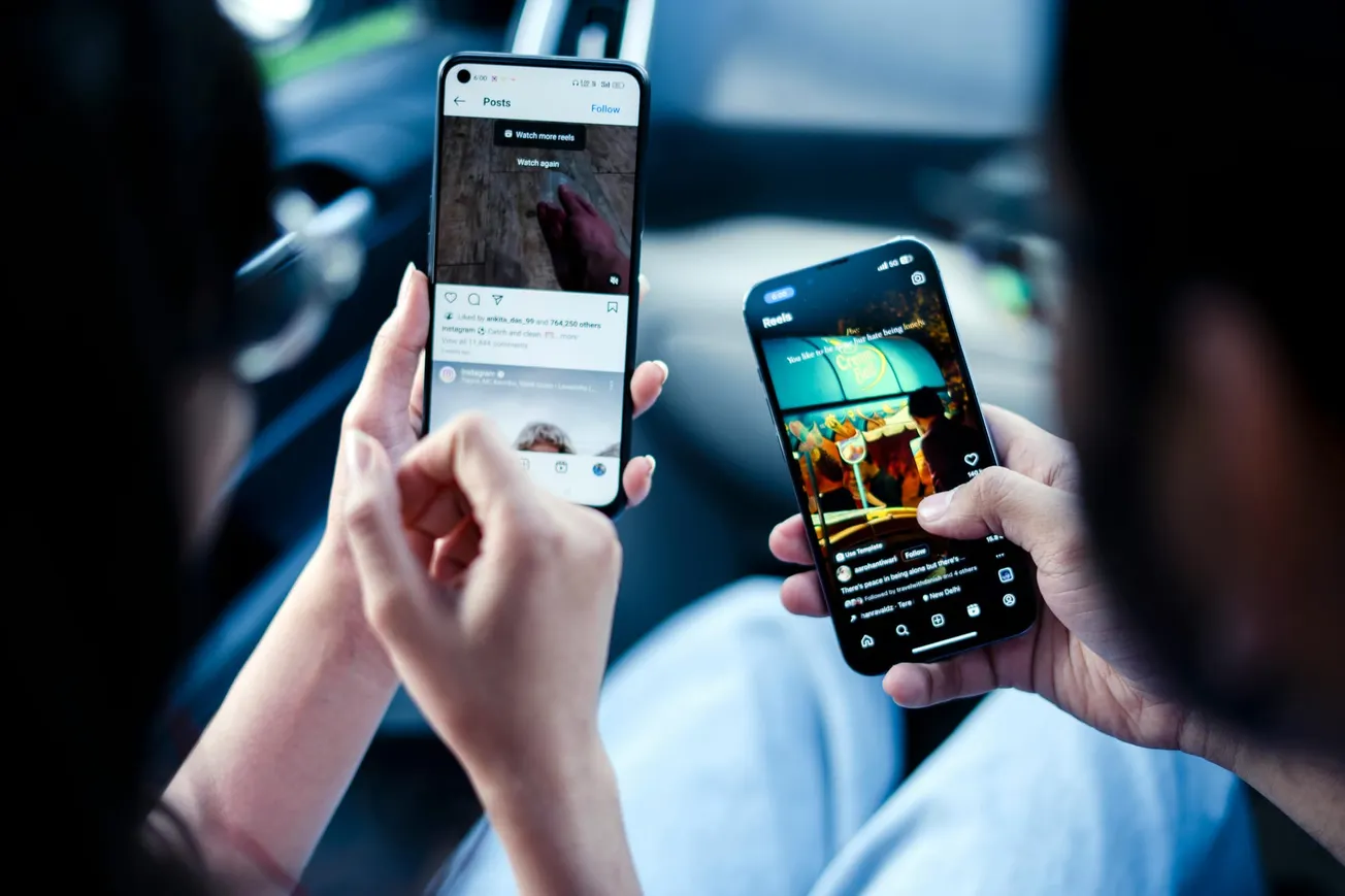 Two people sit closely in a car, each holding a smartphone. Both screens show social media apps.
