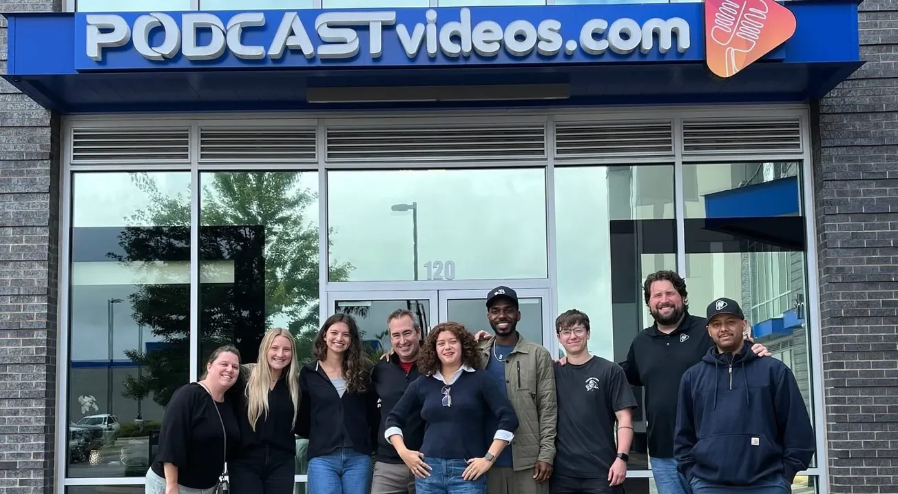 A group of nine smiling people stands in front of a building with the sign "PODCASTvideos.com." The mood is friendly and welcoming.