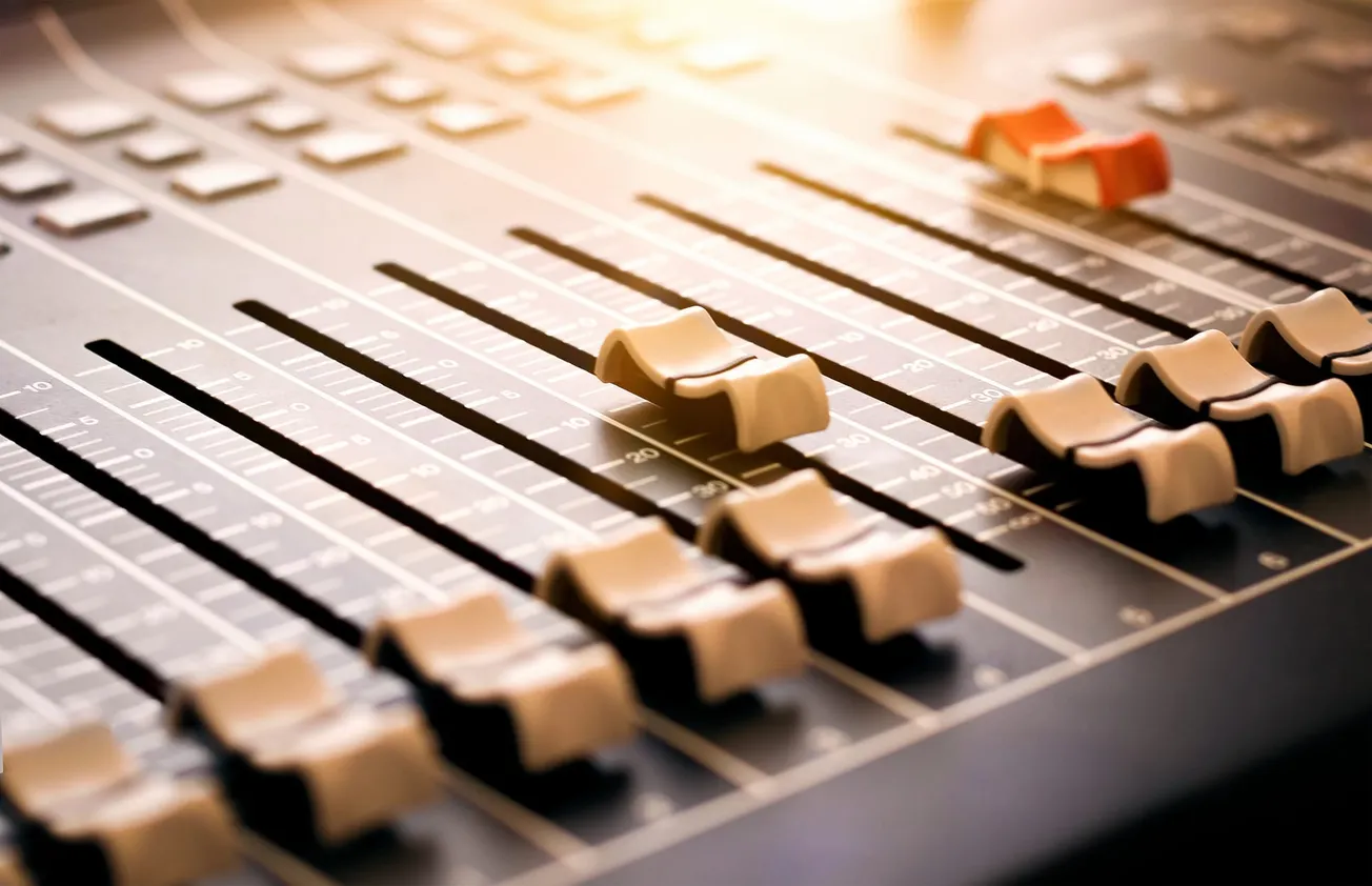 A close-up of a sound mixing console with various beige and one red slider. Soft, warm lighting creates a focused, professional atmosphere on audio control equipment.