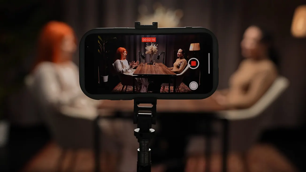 A smartphone on a tripod records two people seated at a table, engaged in a conversation. The setting is cozy and dimly lit, conveying a focused, intimate atmosphere.