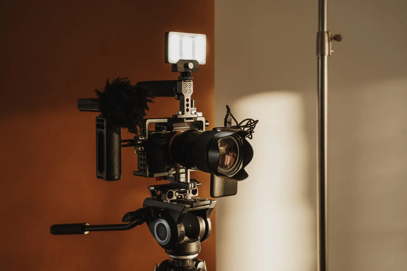 A professional video camera on a tripod, equipped with a microphone and LED light, is set against a brown background with soft lighting.