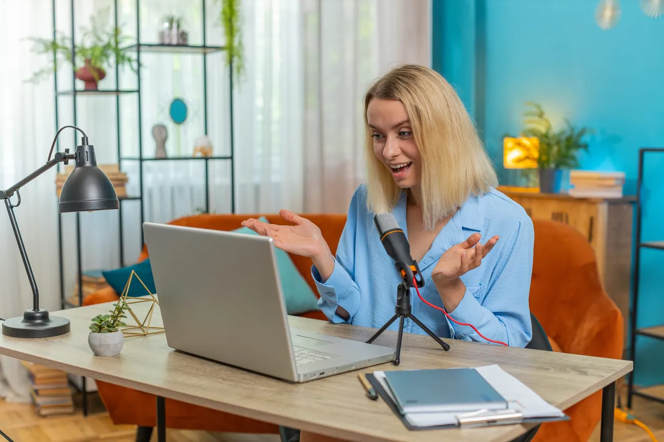 Woman with blonde hair engaging in a video call, smiling and gesturing while seated at a desk with laptop and microphone. Background is a cozy home office.