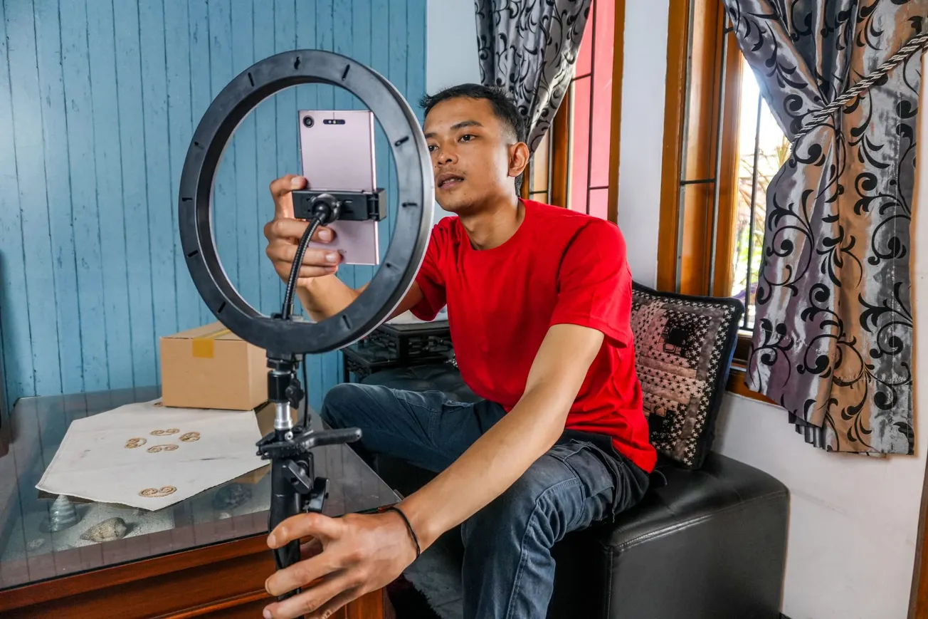 A person in a red shirt sits on a black chair, adjusting a smartphone mounted on a ring light. They're in a cozy room with patterned curtains and a package on a wooden table.