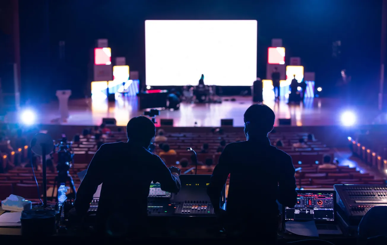 Silhouetted behind sound mixers, two technicians oversee a brightly lit stage with musicians performing, amidst a large audience in a theater.