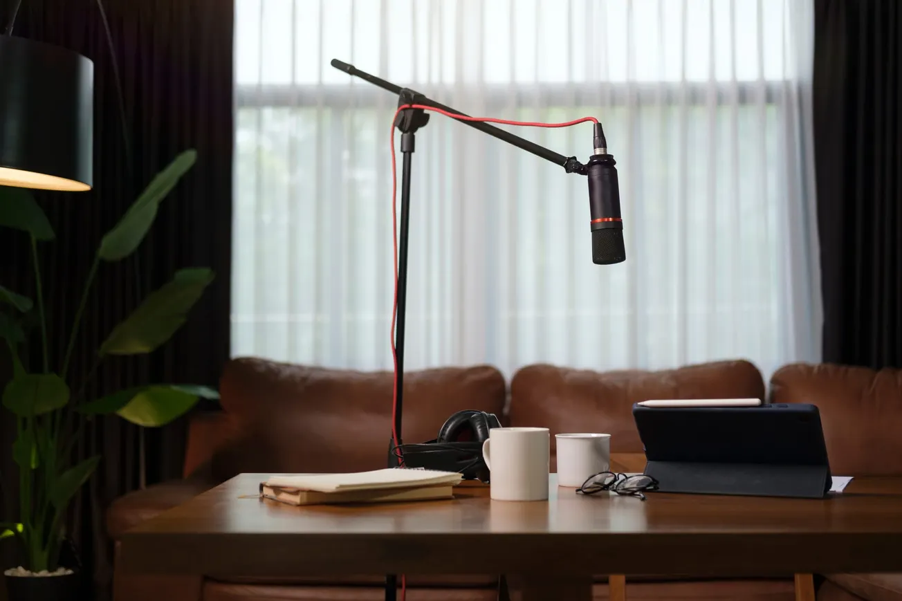 Cozy podcast setup with a microphone on a stand, tablet, and notebooks on a table in front of a brown sofa. Soft lighting and a potted plant create a relaxed atmosphere.