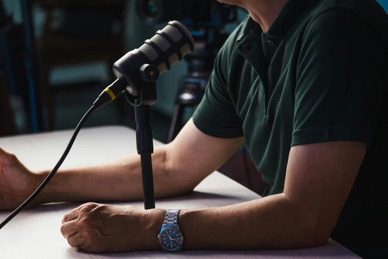 A person in a green shirt is speaking into a microphone on a desk, with a focus on their arm and a wristwatch. A camera is blurred in the background.