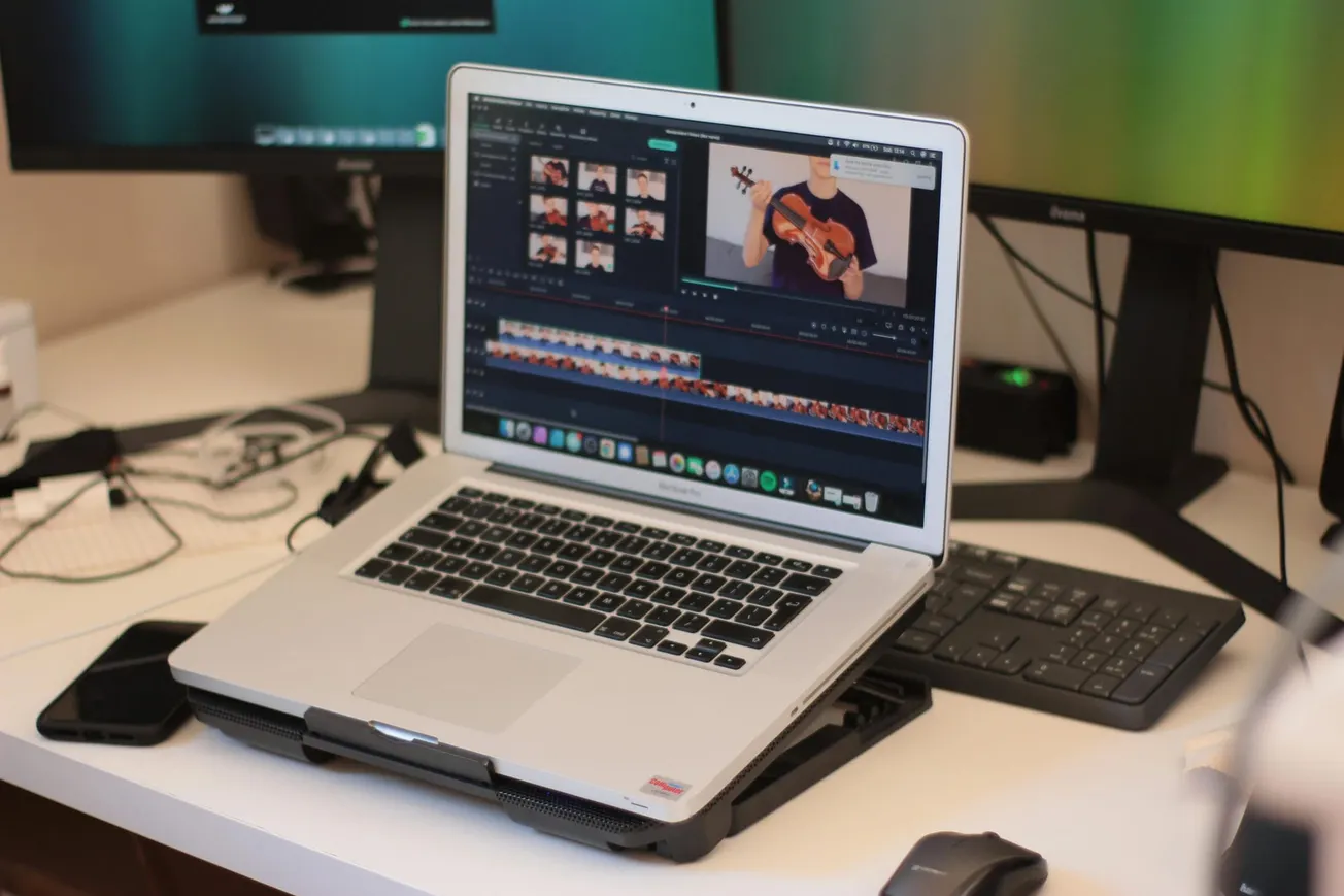 A laptop on a desk shows video editing software, featuring a person playing a violin. Nearby are a keyboard, mouse, and smartphone.