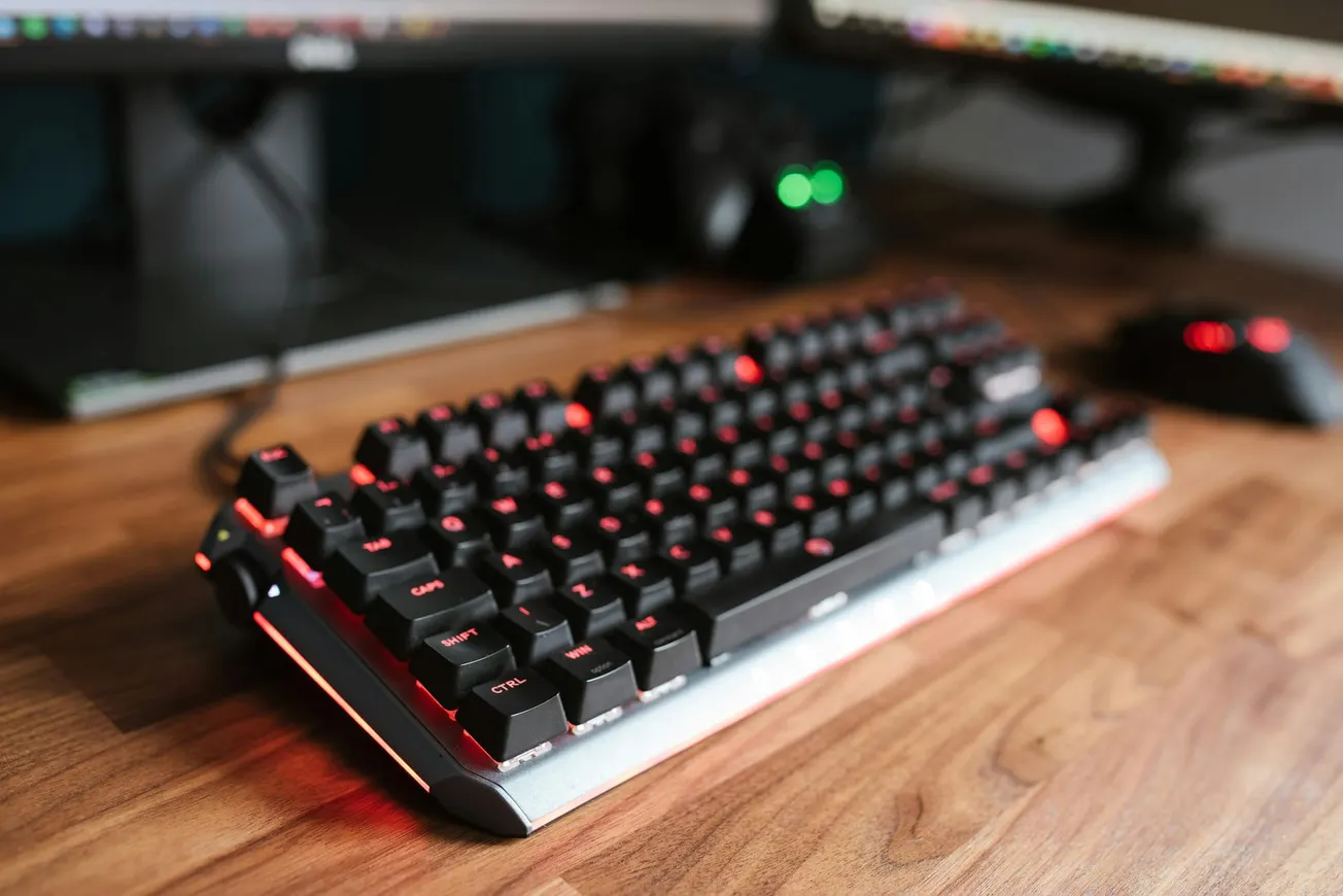 Mechanical gaming keyboard with red backlit keys on a wooden desk, next to a gaming mouse and dual monitors.