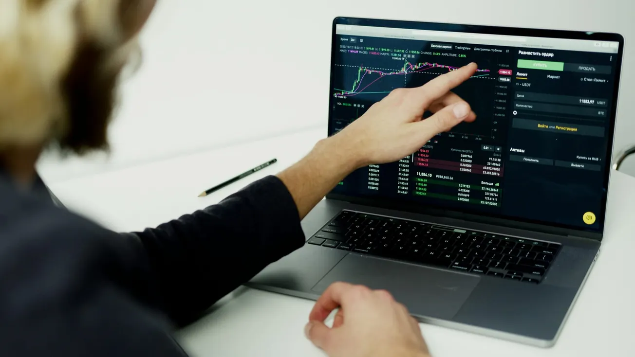 A person points at a laptop screen displaying stock market charts and data. A pencil lies nearby on the white desk.