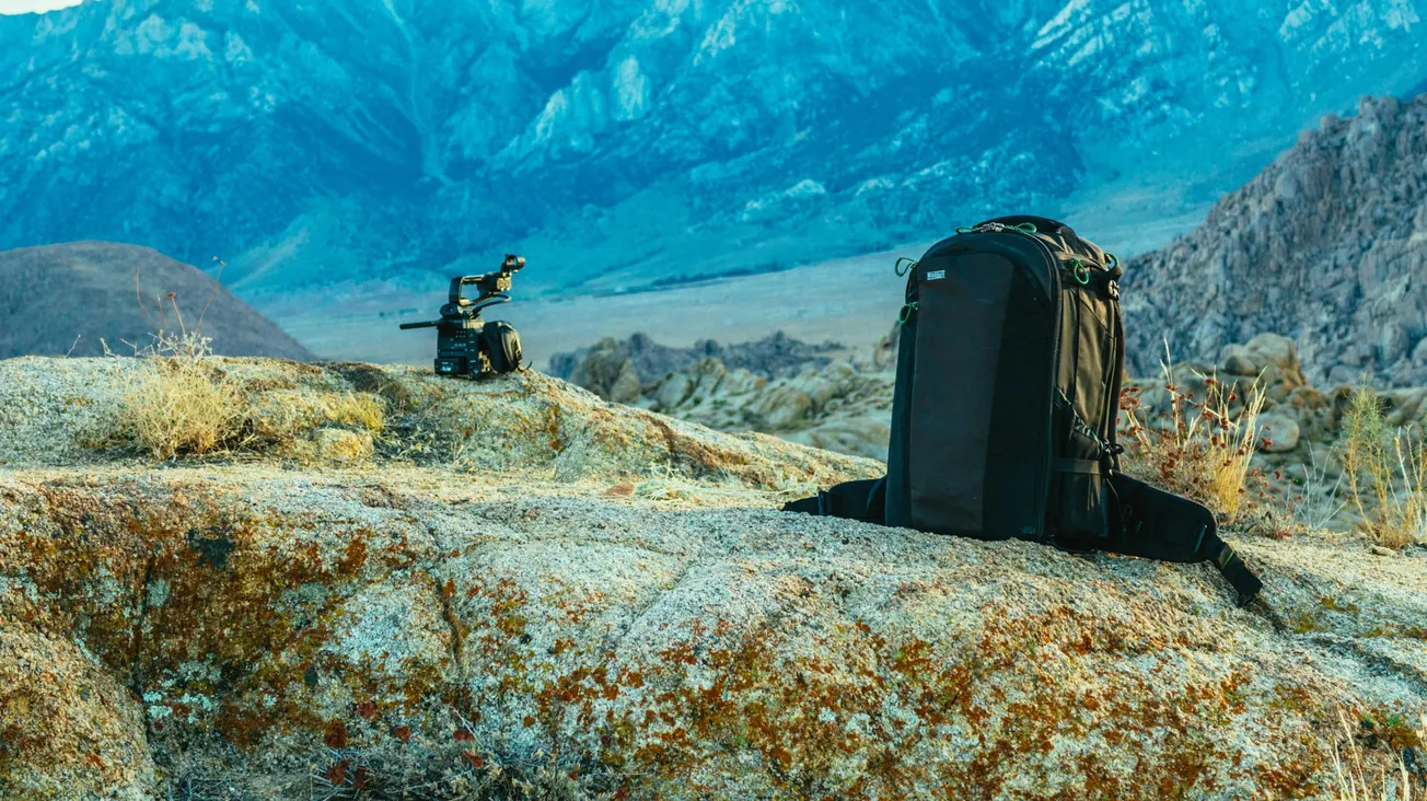 Backpack and camera on rocky terrain with rugged mountains in the background. The scene conveys adventure and exploration under a clear sky.