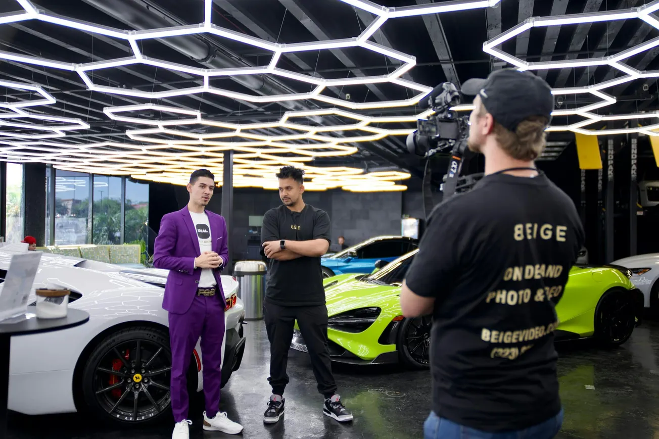 Two men stand in a modern showroom with luxury cars. Another man holds a camera, videoing them talking. One onscreen wears a purple suit, the other casual black. 