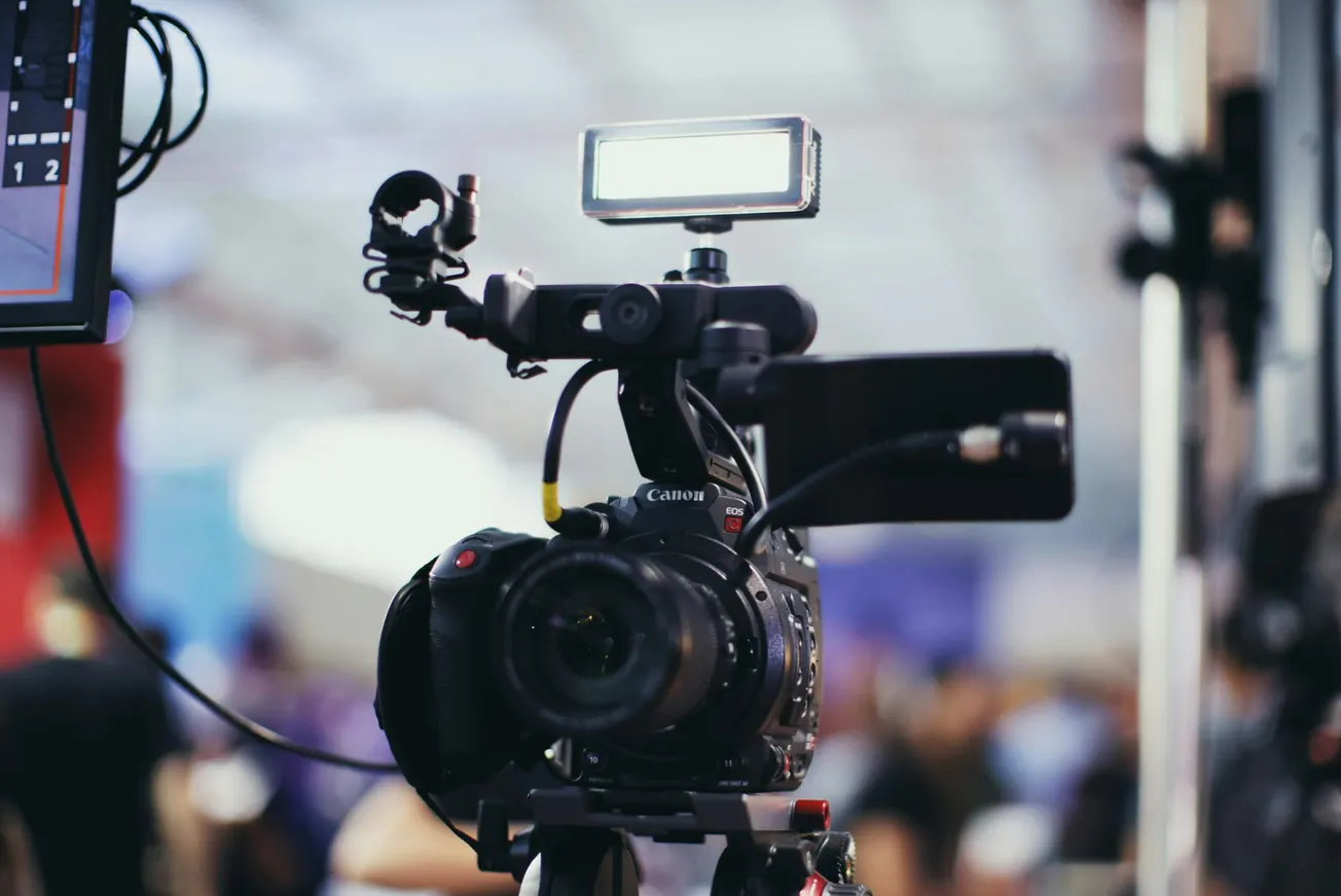 Close-up of a Canon video camera with an attached light and microphone in a studio setting. The blurred background indicates a busy, professional environment.