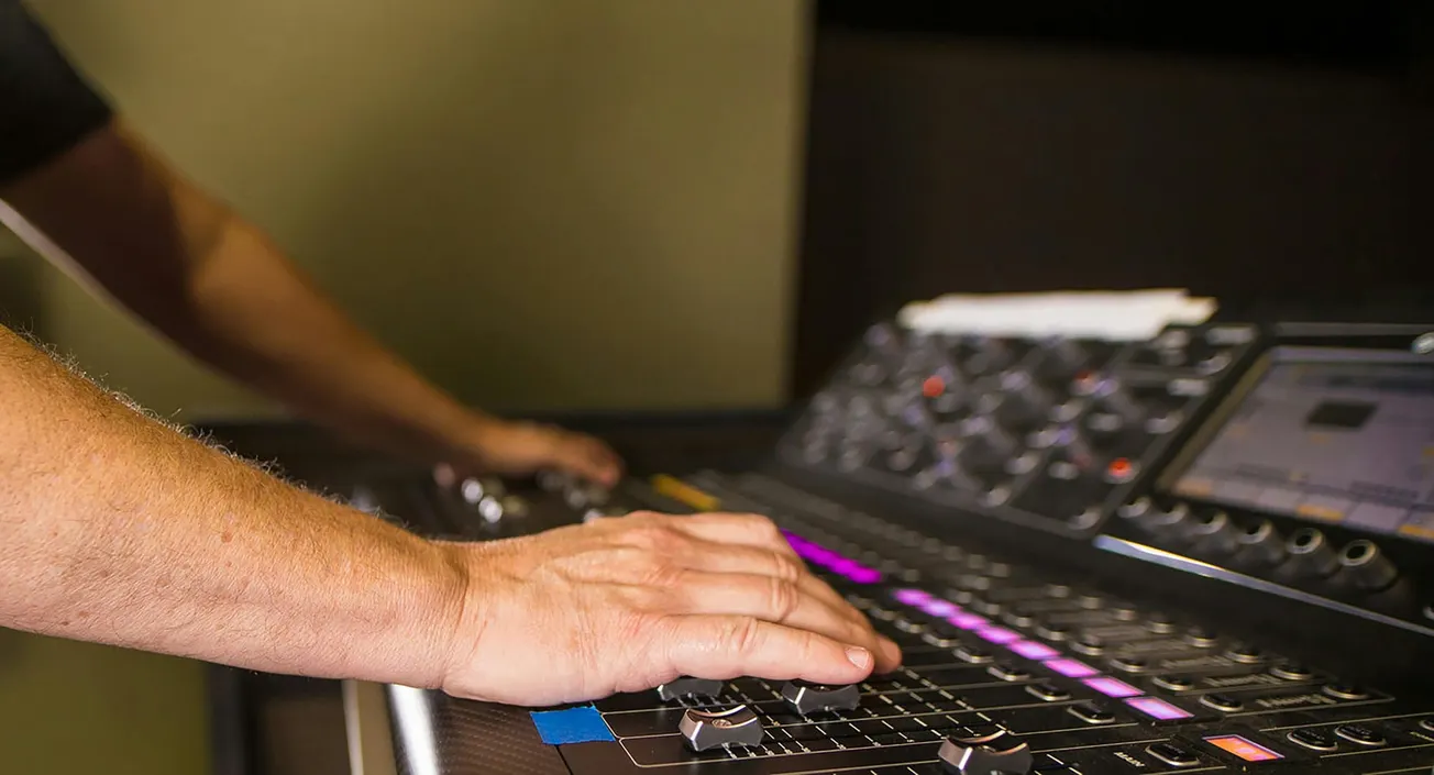 Hands adjusting controls on a professional audio mixing console. The colorful LED lights on the panel indicate an active session, creating a focused atmosphere.