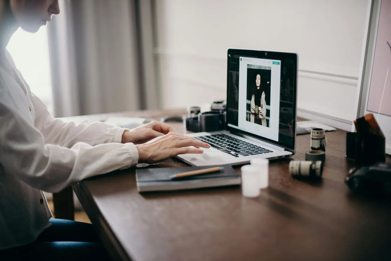A person is typing on a laptop at a wooden desk, surrounded by camera lenses and film canisters. 