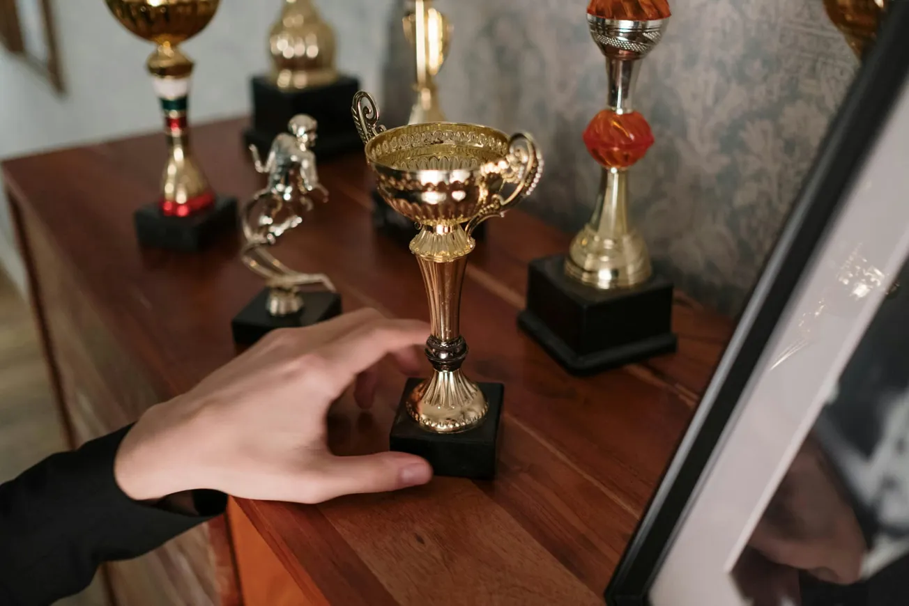 A hand gently grasps a gold trophy among several awards on a wooden shelf. A photo frame is partially visible.