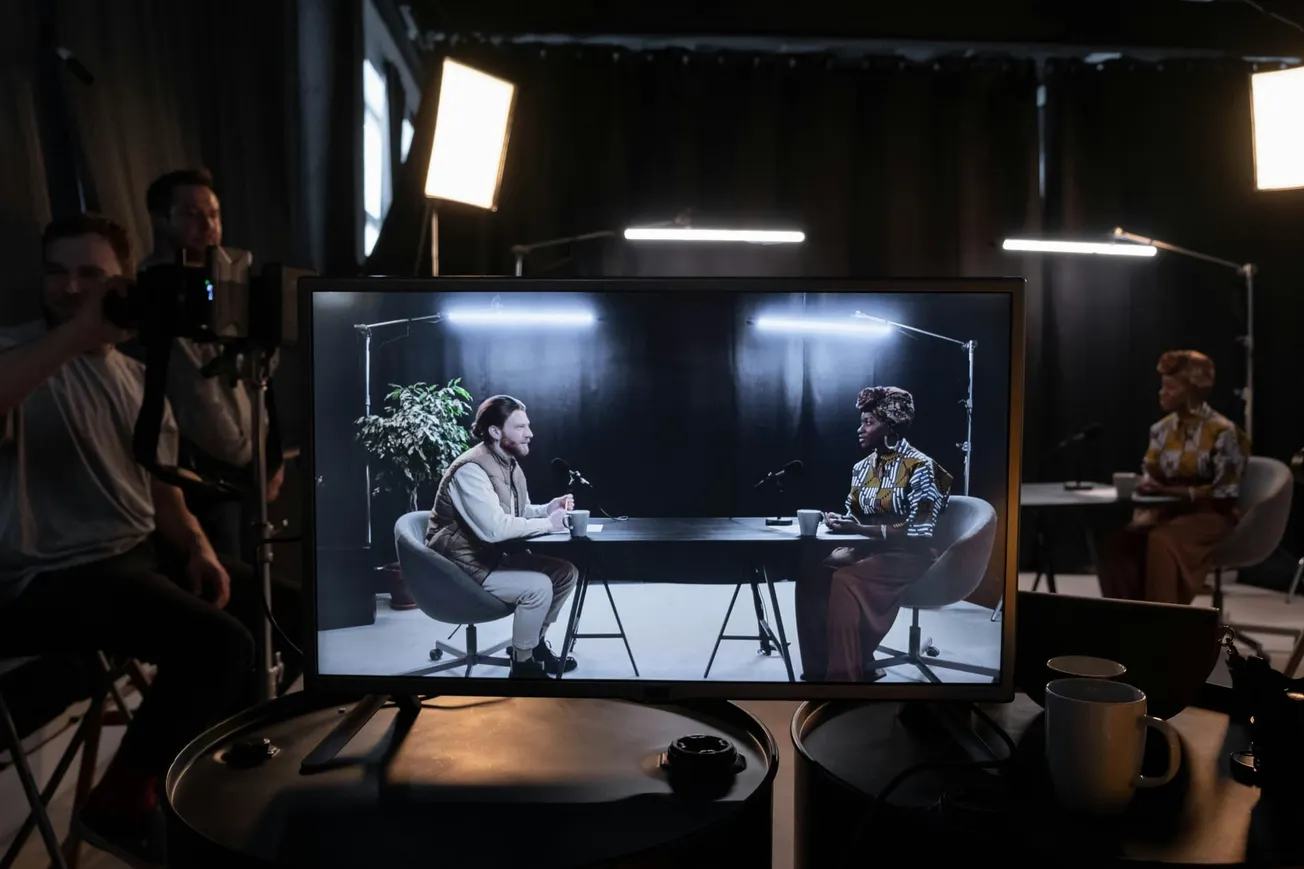 A podcast recording setup shows two hosts seated at a table on a monitor. The studio is dimly lit with bright lights focusing on them.