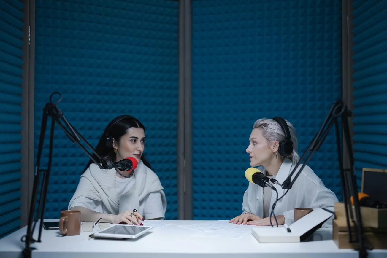 Two women seated in a blue soundproof studio, engaged in conversation. Each wears headphones and speaks into a microphone, creating a focused, professional atmosphere.