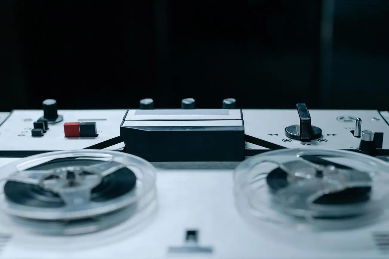 Close-up of a vintage tape recorder with visible reels and control knobs, set against a dark background. The scene conveys a nostalgic, retro vibe.