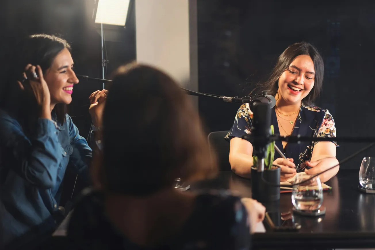 Three women in a podcast studio are laughing and engaged in conversation. Two are wearing headphones, and microphones are placed on a table, creating a lively and friendly atmosphere.