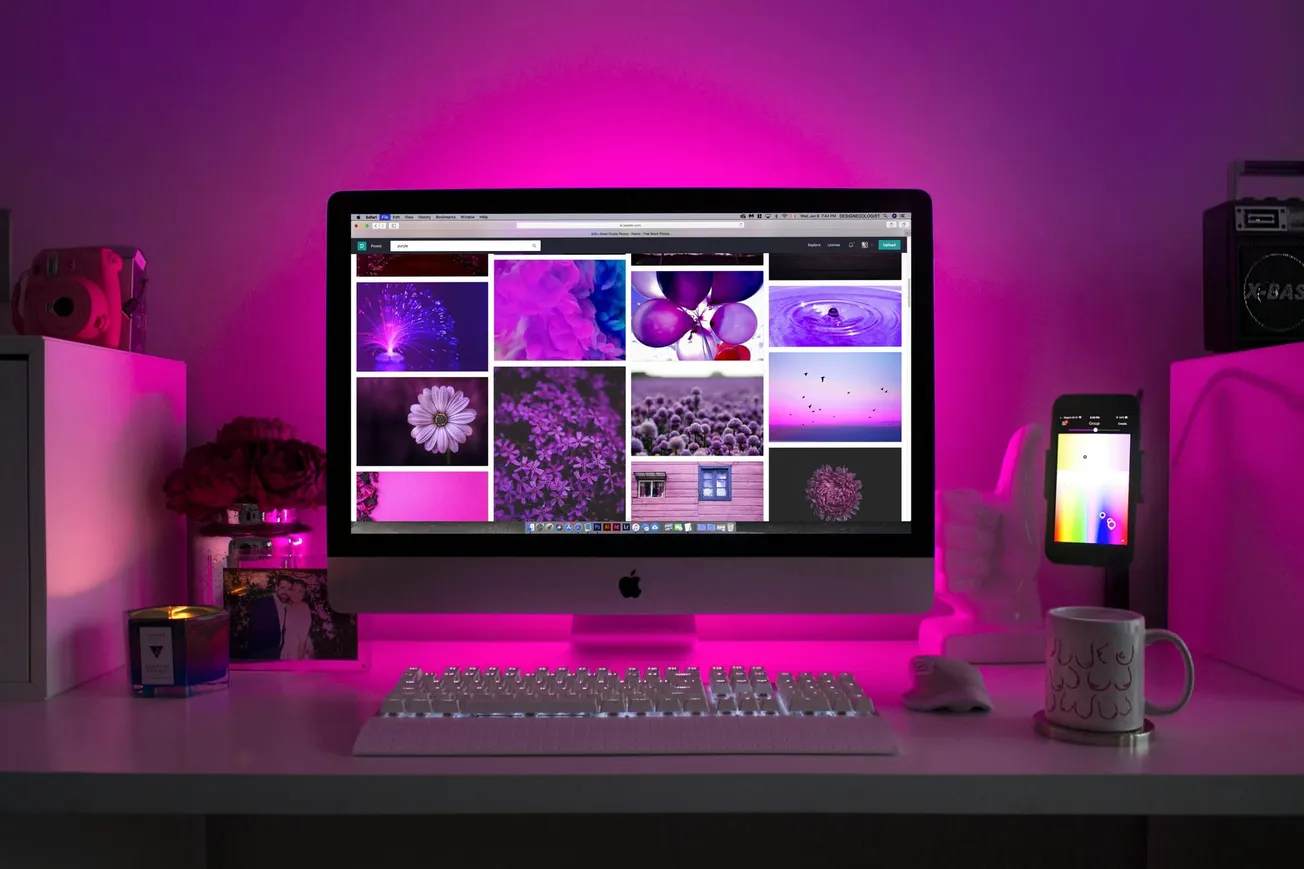 A desktop computer displays purple-themed images under pink ambient lighting. A keyboard, mouse, phone with a color app, candle, and mug adorn the desk.