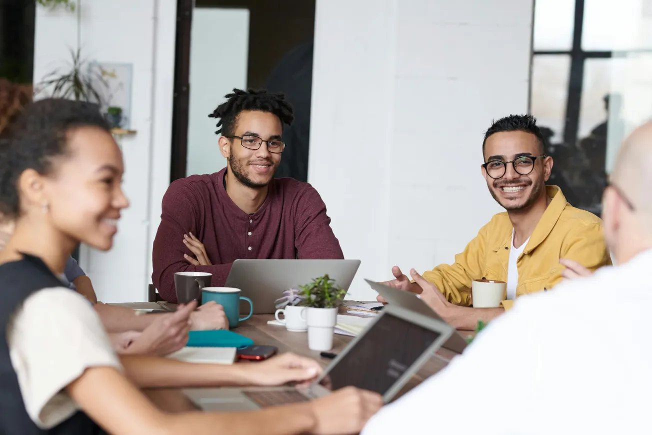 A diverse group of people smiling in a casual meeting setting, seated around a table with laptops and coffee mugs, creating a positive and collaborative atmosphere.