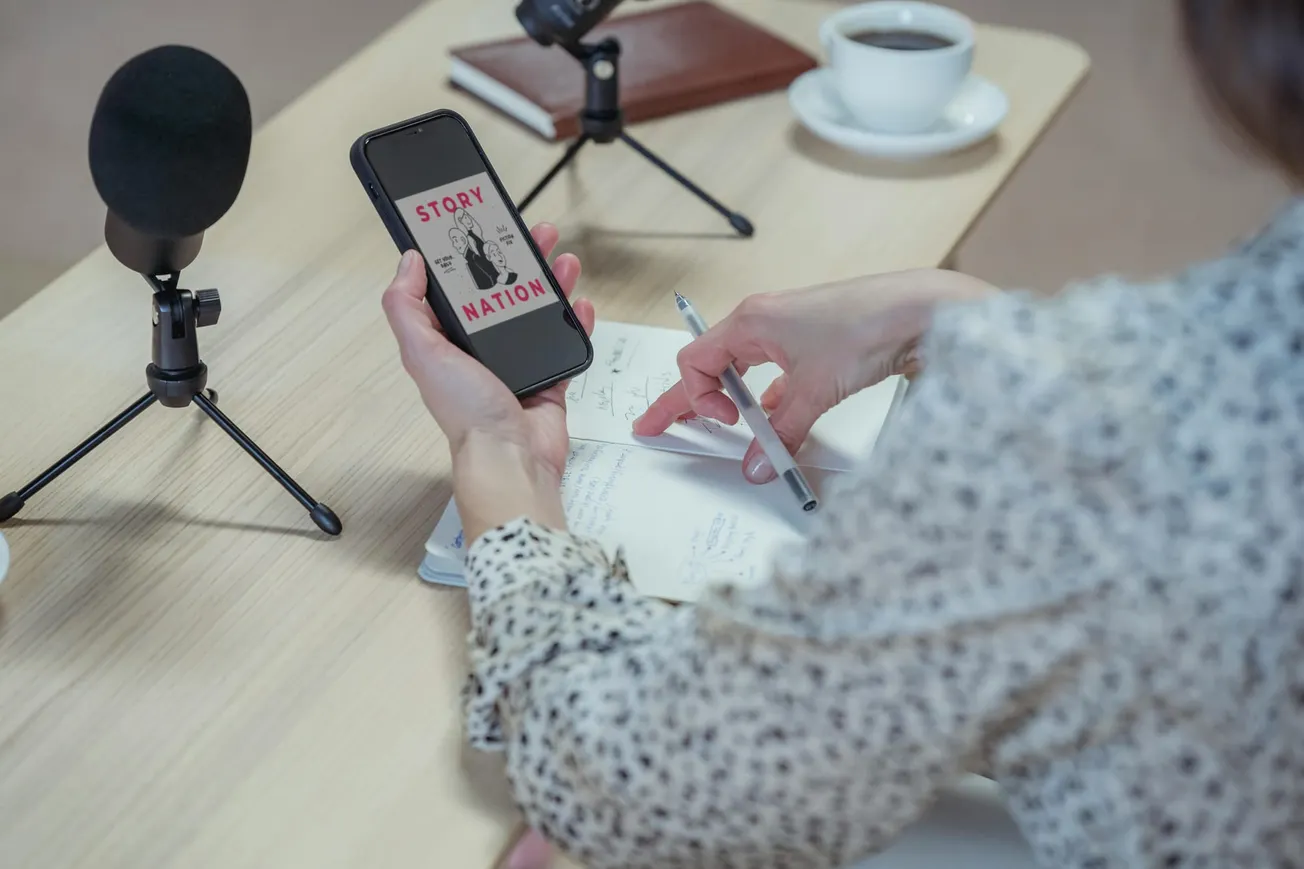 A person sits at a wooden table with a smartphone displaying "Story Nation," taking notes. Two microphones and a coffee cup create a focused podcasting atmosphere.