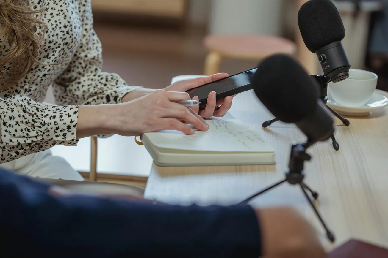 Hands holding a smartphone and writing in a notebook, with microphones on a table. The setting suggests a podcast or interview recording.