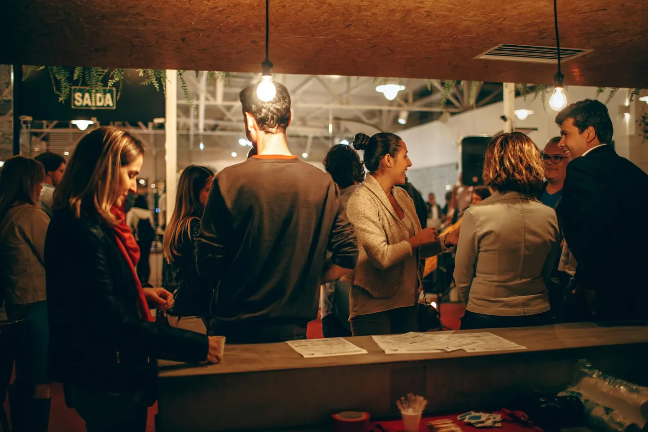 A group of people socializing indoors under warm lighting, creating a cozy atmosphere. They stand around a table with papers, engaged in conversation.