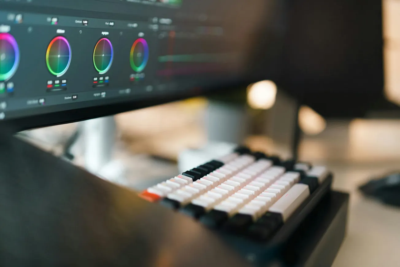 A keyboard in focus with white, black, and orange keys sits in front of a monitor displaying color correction wheels.