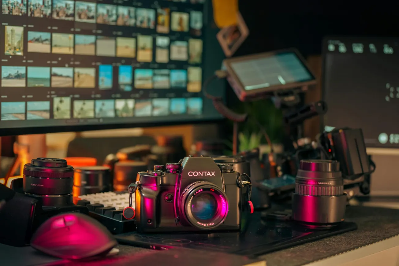 A vintage Contax camera with a pink light glows on a cluttered desk beside a mouse, lenses, and monitors displaying photo thumbnails, creating a nostalgic atmosphere.