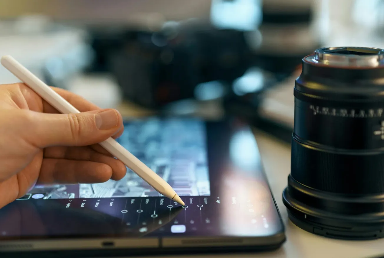 A hand uses a stylus on a tablet displaying photo editing software, next to a camera lens. The scene conveys creativity and focus.