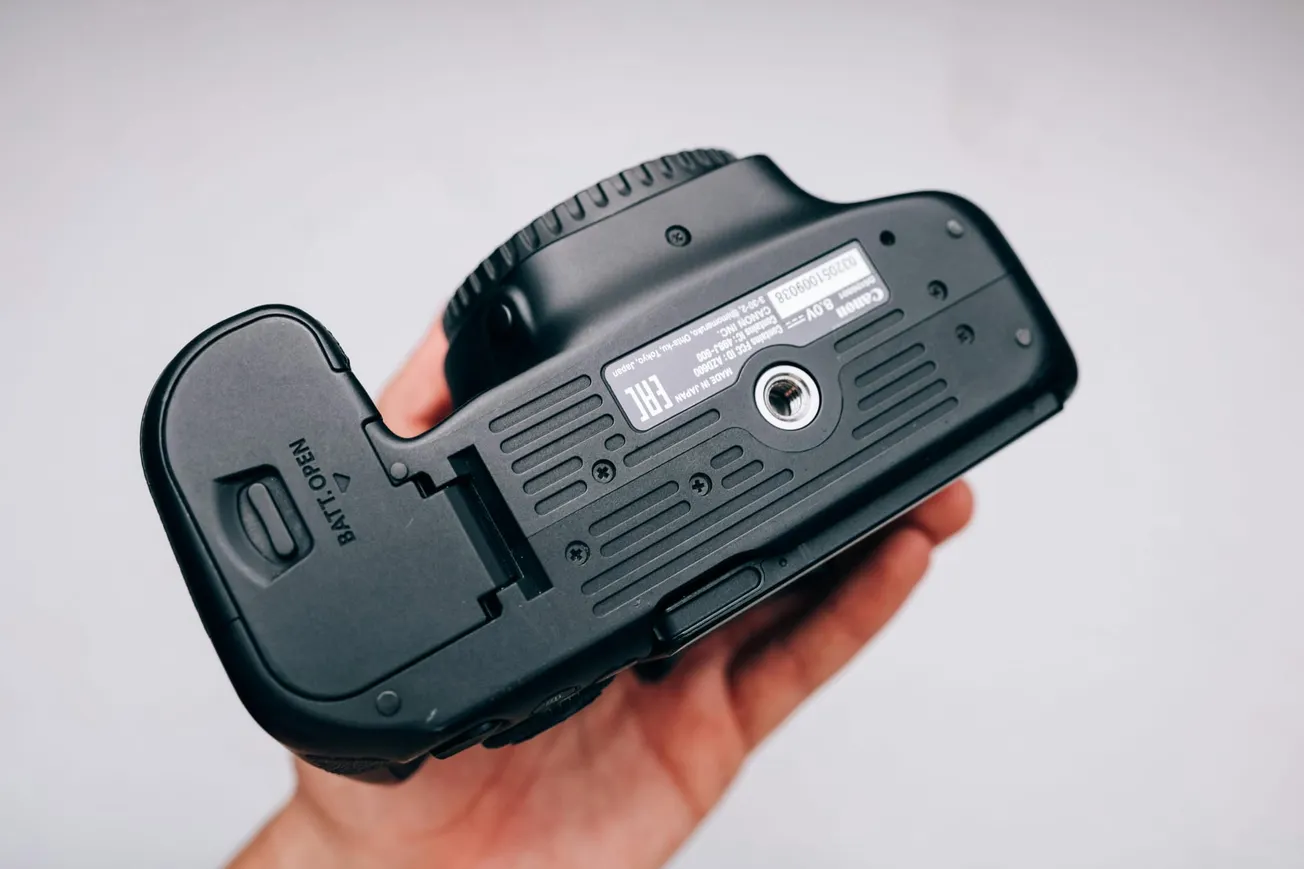 A person holds a camera upside down, showing the bottom with a tripod mount, battery compartment, and informational label.
