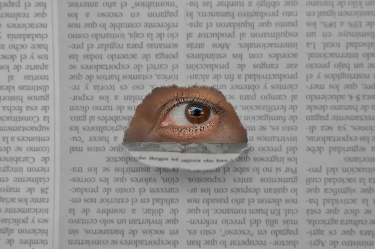 A brown eye peers through a tear in a newspaper, which is displayed upside down. The eye conveys a sense of curiosity or intrigue against the text backdrop.