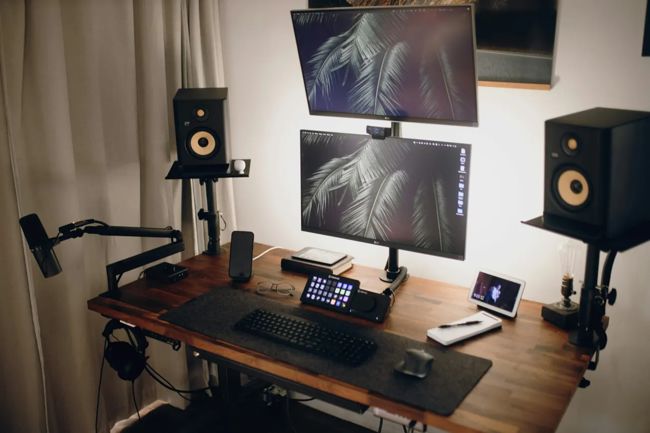 A modern home office desk setup with dual monitors displaying a leafy pattern, flanked by speakers. A microphone, keyboard, tablet, and phone are on the wooden desk.