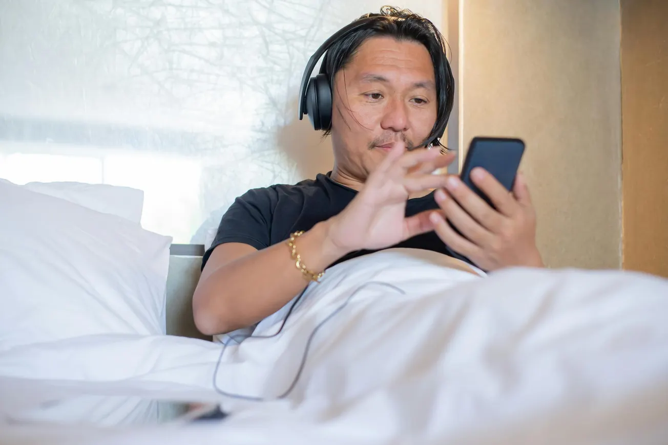 Man in bed wearing headphones, focused on a smartphone. Relaxed atmosphere with white bedding. Casual and calm setting, suggesting leisure time.