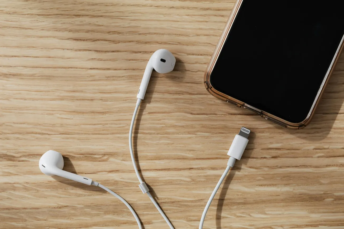 White earphones with a Lightning connector placed beside a smartphone on a light wooden surface.