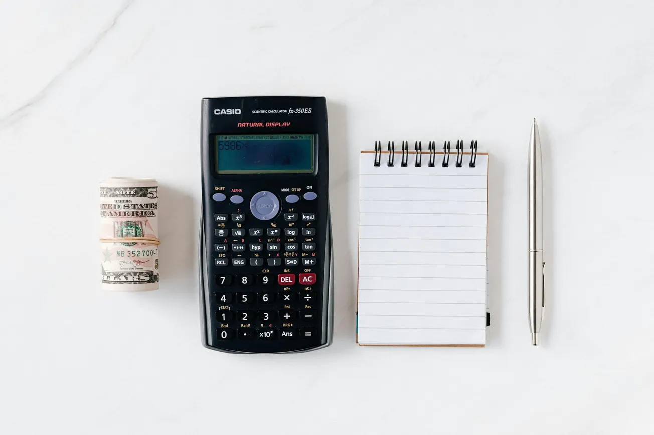 A scientific calculator, a spiral notepad, and a pen are lined up on a white surface. A roll of US dollar bills is placed beside the calculator. 
